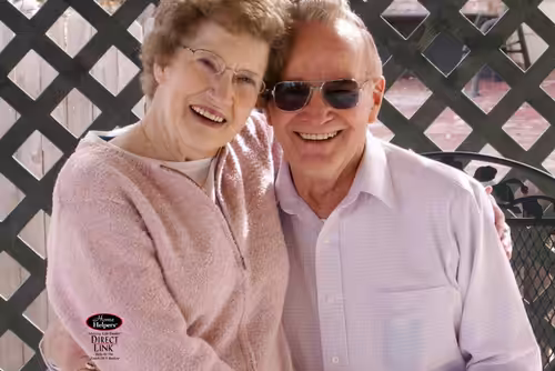 An elderly couple smiles and poses together outdoors, sitting closely with arms around each other in front of a lattice fence. The woman wears glasses and a light pink sweater, while the man wears sunglasses and a light shirt.