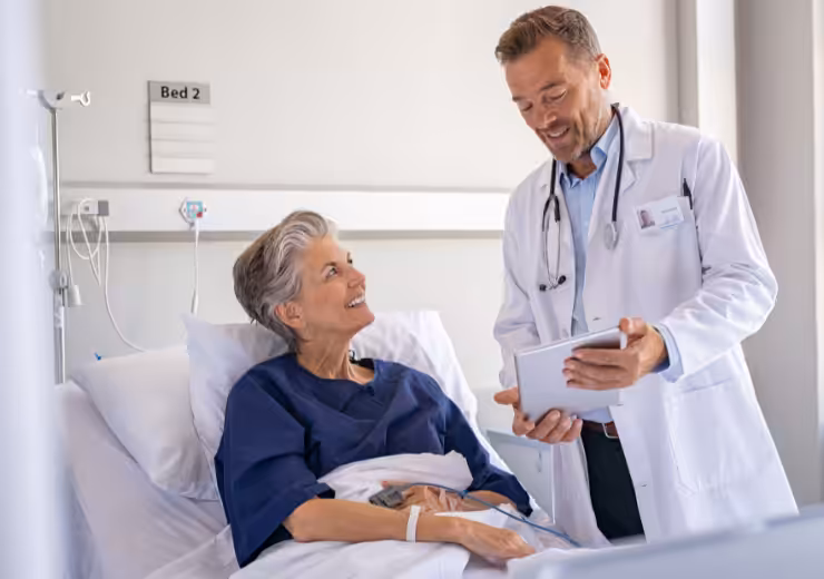 An older woman in a hospital bed smiles and talks with a male doctor holding a tablet. They appear to be having a friendly conversation in a bright hospital room labeled Bed 2.