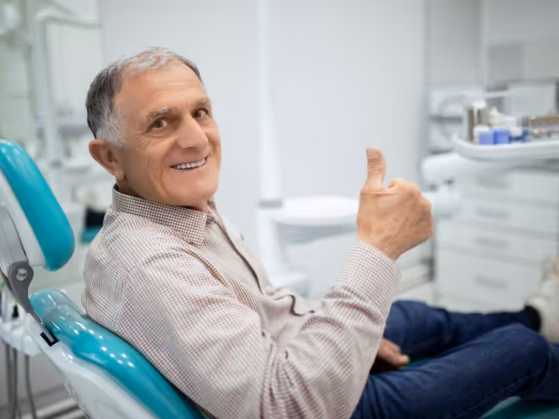An older man sitting in a dental chair smiles and gives a thumbs up, indicating satisfaction. The background shows dental equipment in a bright, modern clinic.