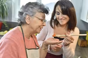 An older woman in glasses and an apron tastes food from a wooden spoon held by a younger woman, both smiling in a kitchen.