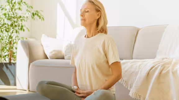A woman sits cross-legged on the floor in a bright living room, eyes closed and hands resting on her lap, appearing calm and relaxed as she meditates. A sofa and blanket are visible in the background.