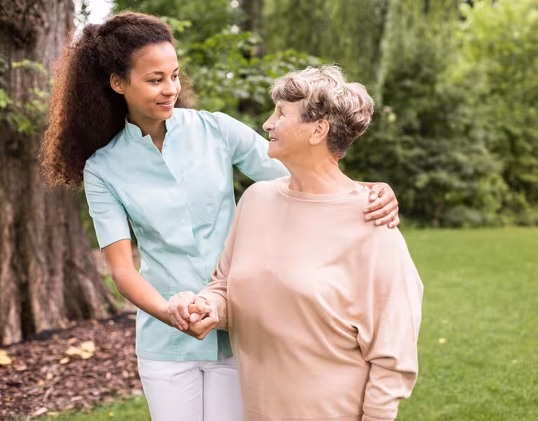 A young caregiver smiles and holds hands with an elderly woman as they stand together outdoors in a green, park-like setting. Both appear happy and connected, sharing a supportive moment.