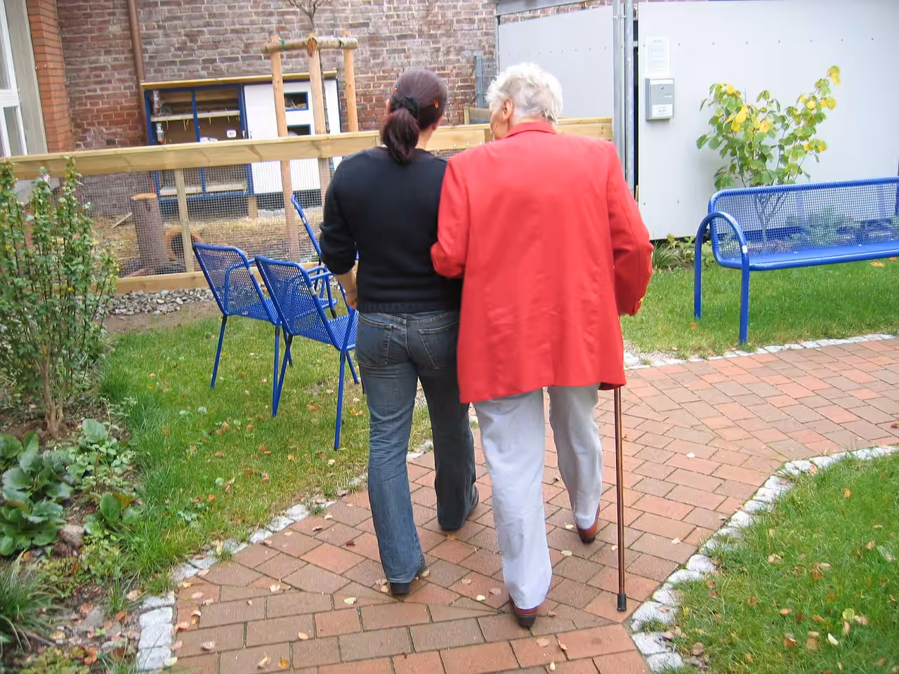 An elderly person with white hair and a cane walks arm-in-arm with a younger person along a brick path in a garden, with blue benches and green grass around them.