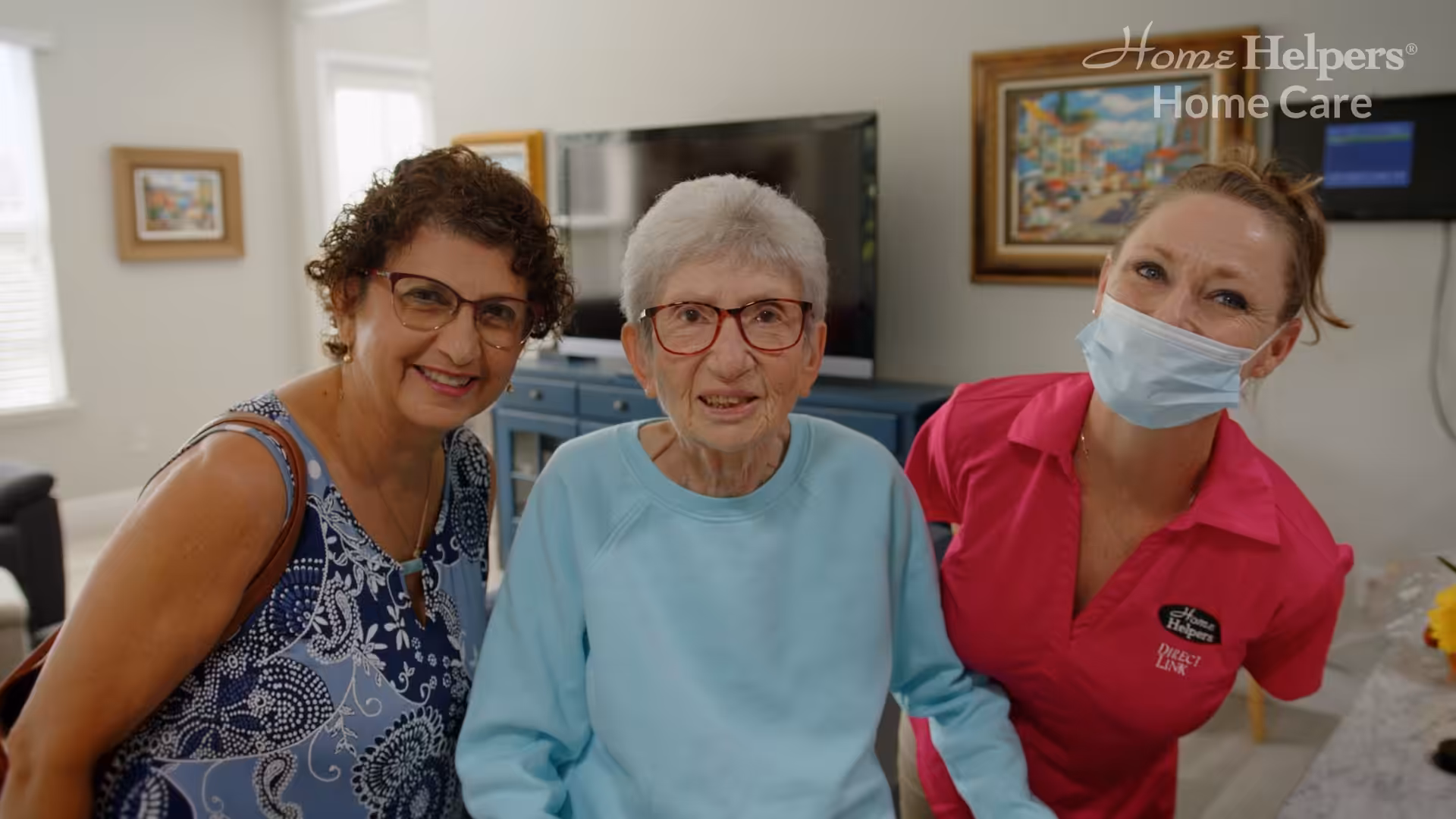 Three women smile together indoors. The woman in the middle has white hair and glasses, the woman on the left has curly hair and glasses, and the woman on the right wears a pink shirt and a face mask. Home Helpers Home Care logo is visible.
