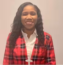 A woman with long curly hair smiles at the camera. She is wearing a red plaid jacket over a white collared shirt and standing in front of a plain, light-colored background.