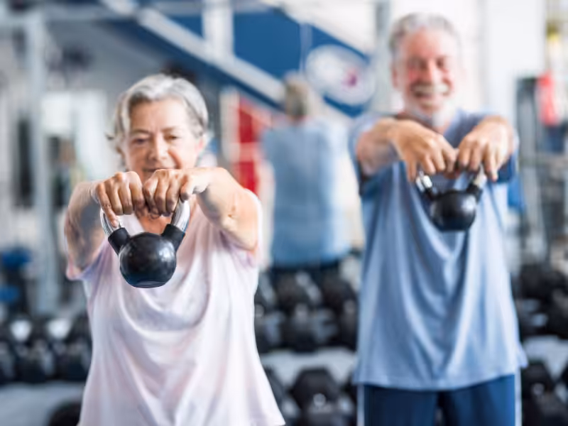 Two older adults exercise in a gym, smiling and lifting kettlebells with both hands, arms extended forward. The background shows gym equipment and weights.