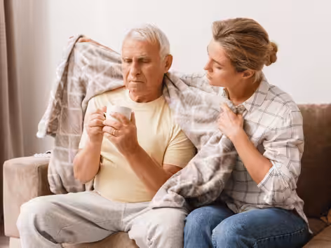 A young woman drapes a blanket over an older man sitting on a couch while he holds a cup, both with serious expressions, suggesting care and support in a home setting.