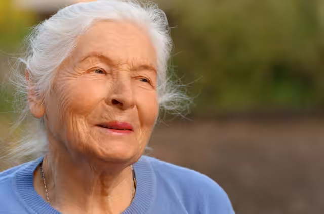 An elderly woman with long white hair and a gentle smile looks slightly to the side. She is wearing a light blue sweater and is outdoors, with a blurred green background.
