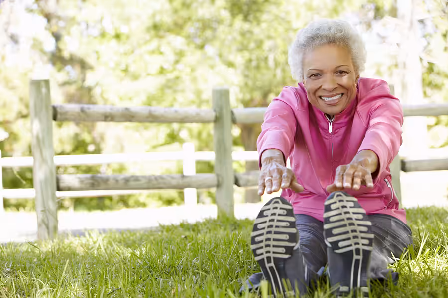 An older woman in a pink jacket sits on grass, smiling as she stretches forward to touch her toes. There is a wooden fence and trees in the background on a sunny day.