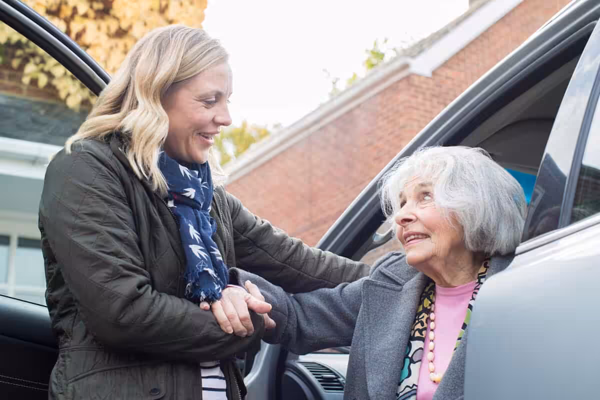 A younger woman smiles and helps an older woman out of a car. The older woman, with gray hair, looks up warmly at the younger woman. They are outdoors near a brick building.
