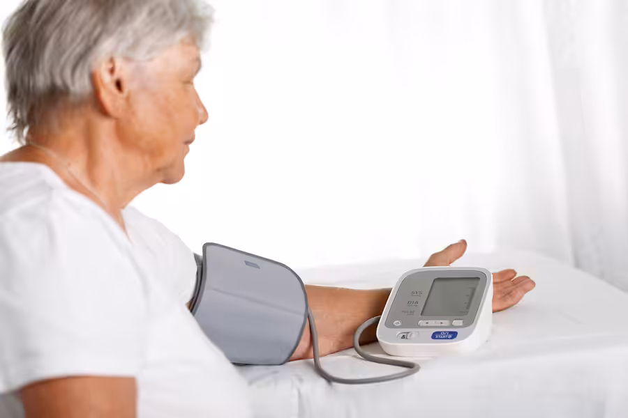 An older woman sits at a table using an automatic blood pressure monitor on her left arm, looking at the digital display as she checks her blood pressure.