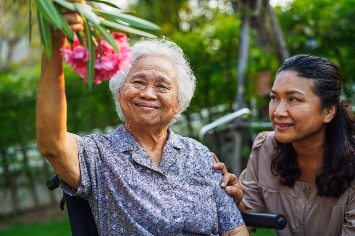 An elderly woman in a wheelchair smiles and holds up pink flowers, while a younger woman beside her gently holds her shoulder and looks at her with affection in a garden setting.