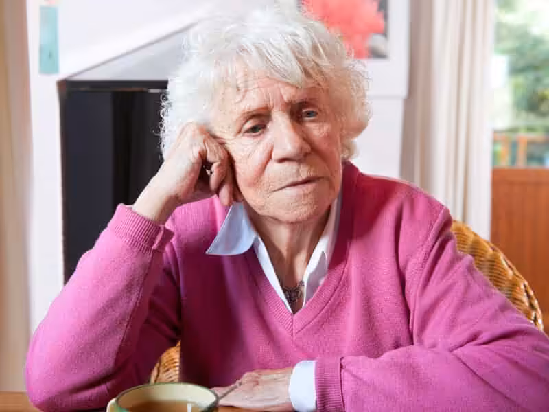 An elderly person with white hair, wearing a bright pink sweater over a white collared shirt, sits at a table resting their head on their hand, looking thoughtful or concerned. A mug is on the table in front of them.
