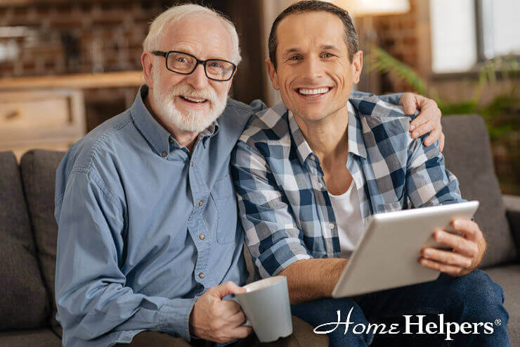 An older man and a younger man sit together on a couch, smiling at the camera. The older man holds a mug, and the younger man holds a tablet. The Home Helpers logo appears in the bottom right corner.