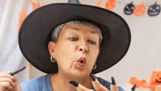 A person wearing a black witch hat and fake long black nails looks at their hand in surprise, with Halloween decorations like pumpkins and ghosts hanging in the background.