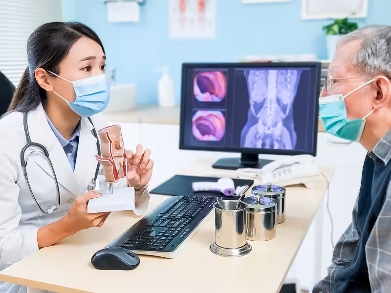 A female doctor wearing a mask explains a medical condition to an older male patient, holding a model of a prostate. X-ray and scan images are displayed on a computer screen in the background.
