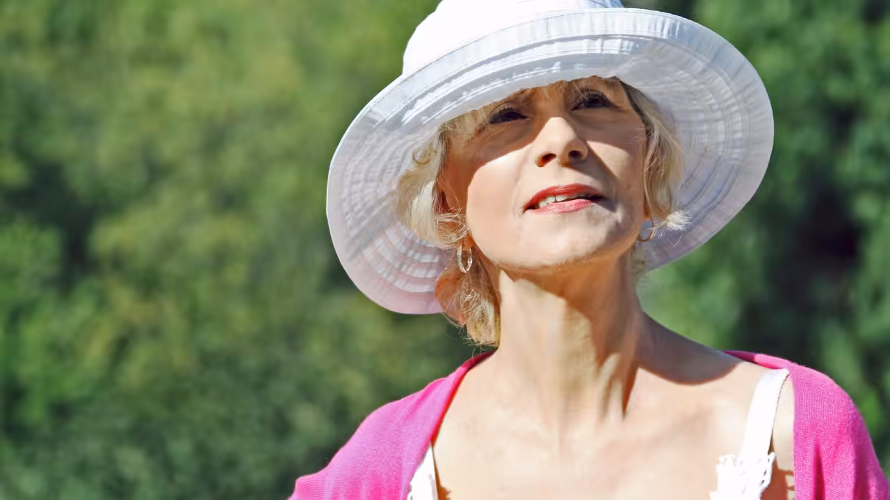 An older woman wearing a wide-brimmed white hat and a pink cardigan stands outdoors, looking up with a gentle smile, against a blurred green background.