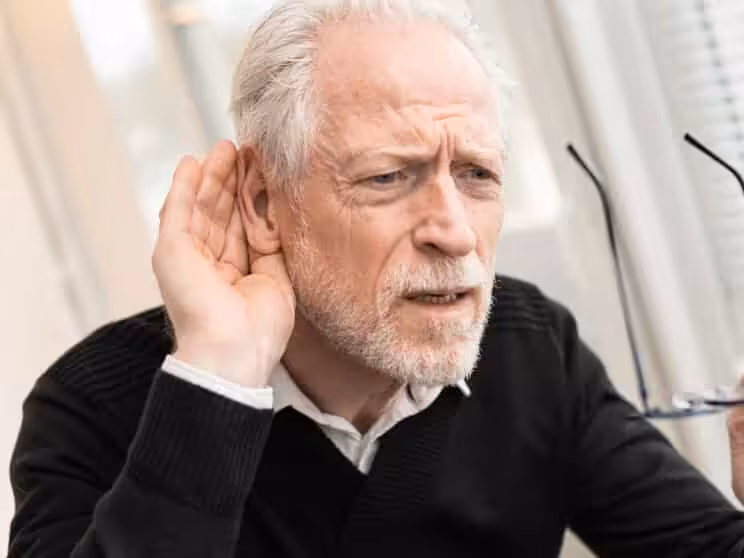 An older man with white hair and a beard leans forward, holding his hand to his ear and squinting as if trying to hear better. He holds a pair of eyeglasses in his other hand.