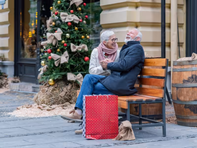 An elderly couple sits on a bench, laughing and holding hands, with a Christmas tree decorated with bows and ornaments behind them. A large red shopping bag rests on the ground in front of them.