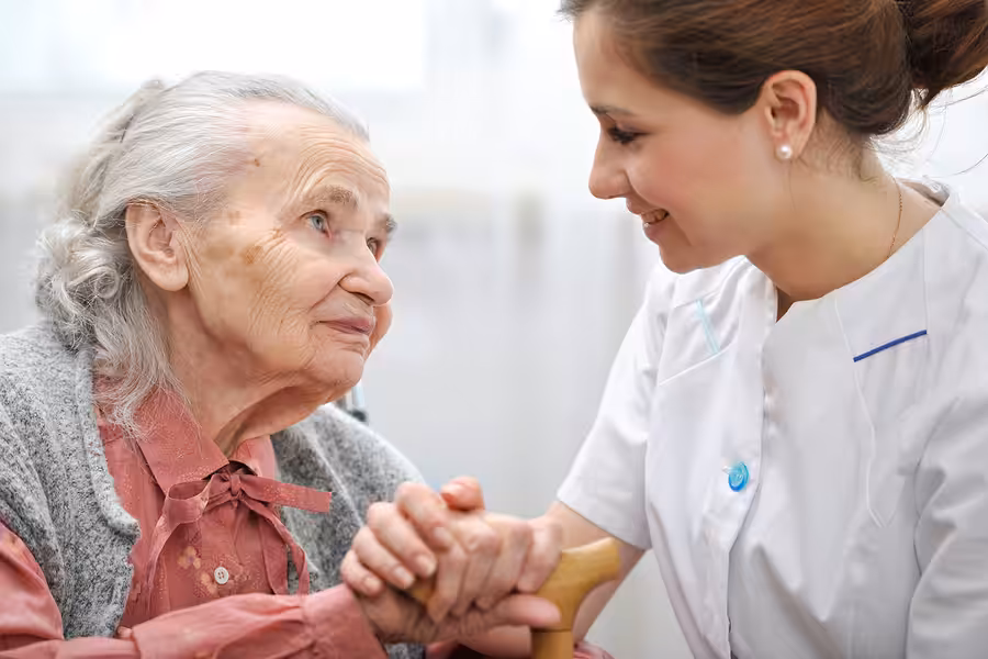 A young female caregiver smiles warmly at an elderly woman, gently holding her hand. The elderly woman looks up at the caregiver with gratitude and affection, creating a moment of connection and support.