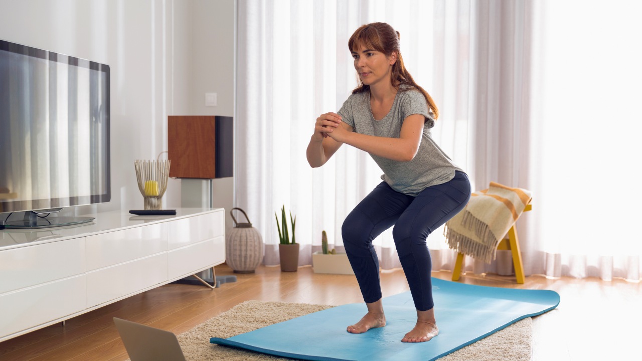 A woman exercising at home stands on a blue mat, performing a squat in front of a TV and a laptop, with a bright living room in the background.