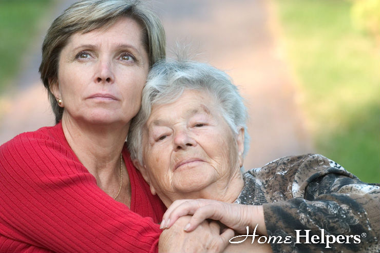 An older woman with gray hair is embraced by a younger woman in a red sweater outdoors. Both look thoughtful. The text Home Helpers is at the bottom right.