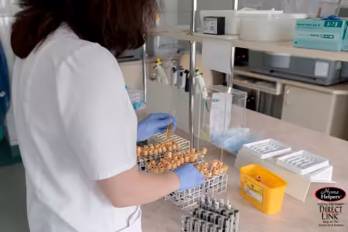 A person wearing a white lab coat and blue gloves organizes blood sample vials in a laboratory setting, with medical equipment and supplies visible on the countertop.