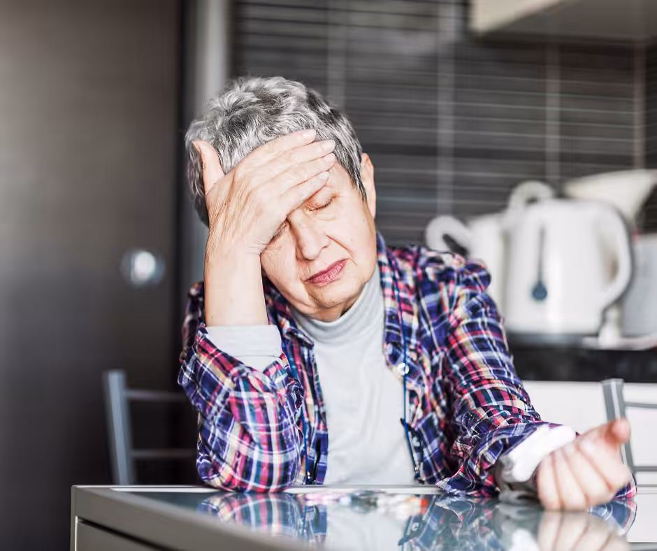 An elderly woman with short gray hair sits at a table, looking tired or unwell, with her eyes closed and one hand on her forehead. She wears a plaid shirt and is in a kitchen setting.