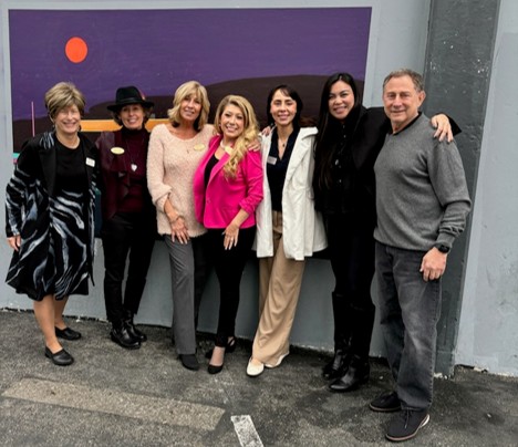 Seven people stand side by side, smiling at the camera in front of a mural featuring a purple sky, hills, and a red sun. They are dressed in casual and business attire.