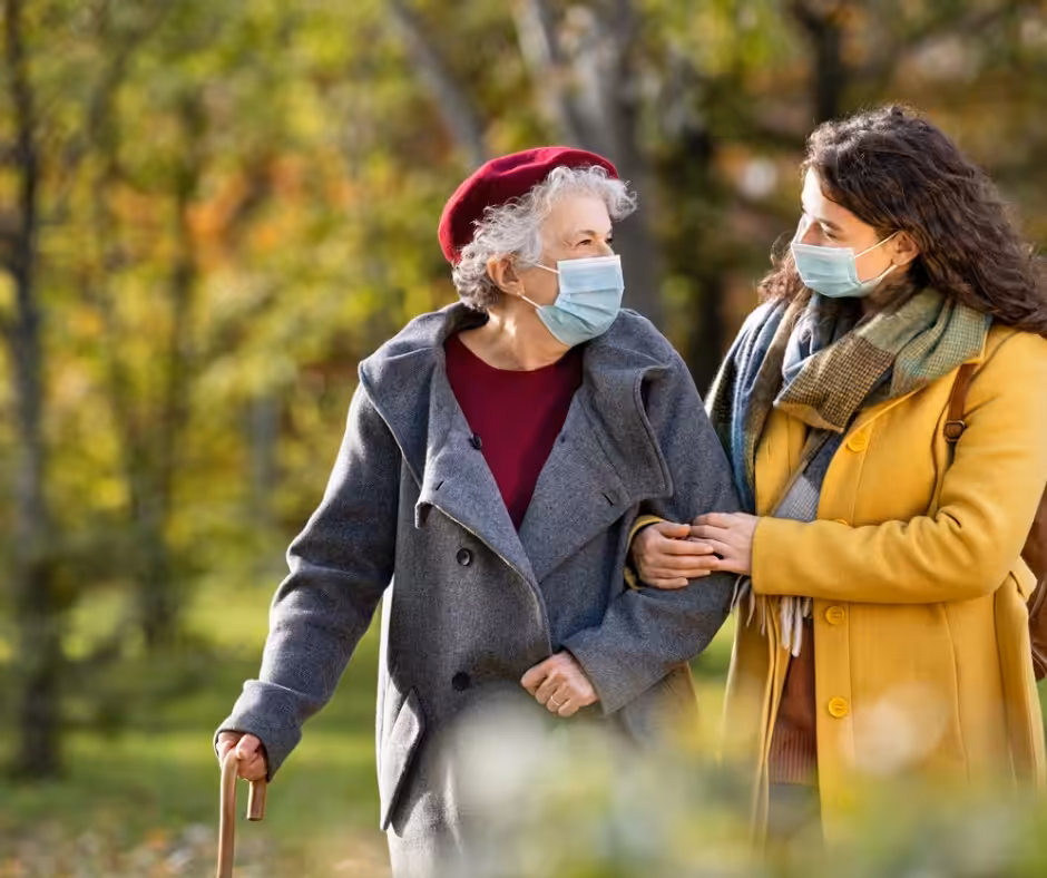 Two women wearing face masks walk arm-in-arm outdoors. One is elderly, using a cane and wearing a red beret and gray coat; the other is younger, wearing a yellow coat and scarf. Trees with autumn foliage are in the background.