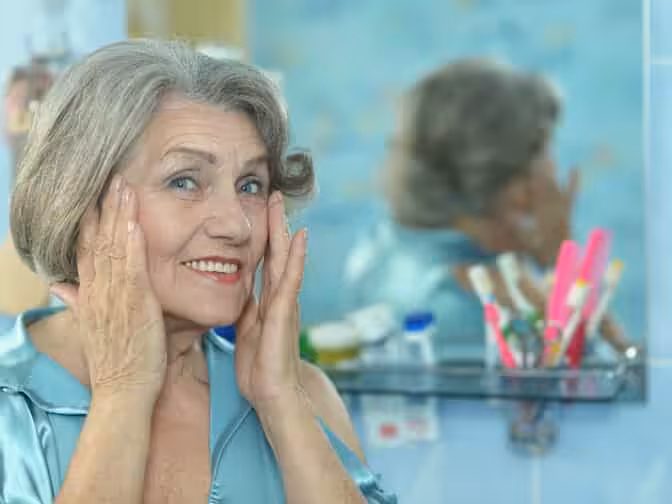 An older woman with gray hair smiles in front of a bathroom mirror, gently touching her face with both hands. Toiletries and toothbrushes are visible on a shelf behind her.
