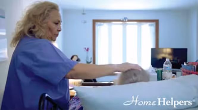 A caregiver in scrubs gently assists a person lying in bed in a bright room. The Home Helpers logo appears in the bottom right corner.