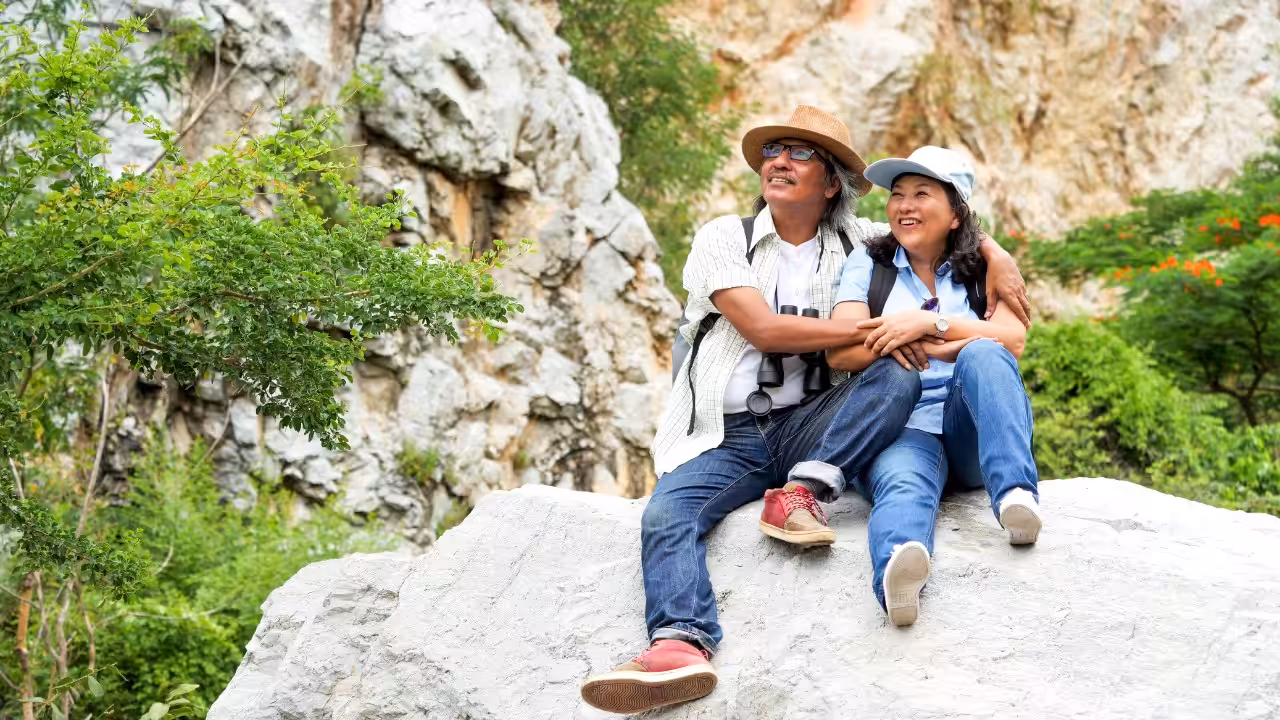 An older couple sits together on a large rock outdoors, smiling and looking into the distance, surrounded by greenery and rocky cliffs.
