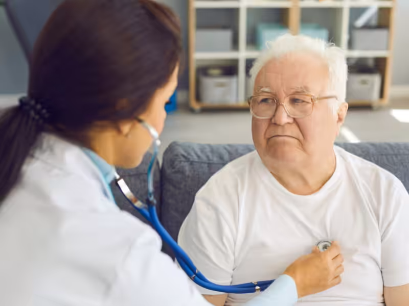 A doctor listens to an elderly mans chest with a stethoscope during a medical checkup at home. The man is wearing glasses and a white t-shirt, and looks concerned as he sits on a couch.