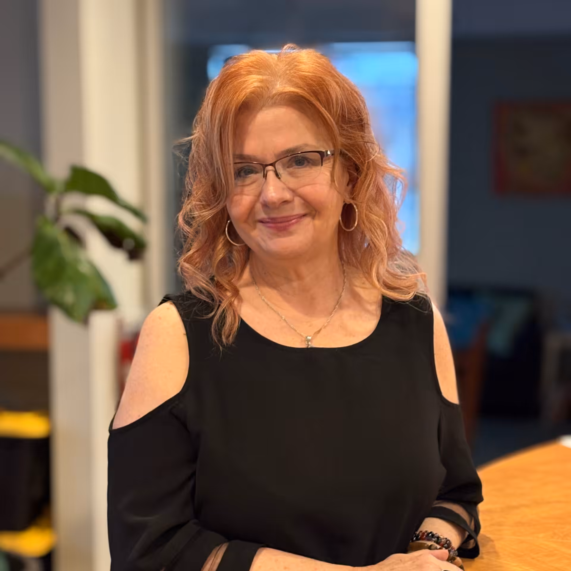 A woman with wavy red hair and glasses, wearing a black cold-shoulder top and hoop earrings, stands indoors smiling at the camera. A large green plant and blurred background furniture are visible.