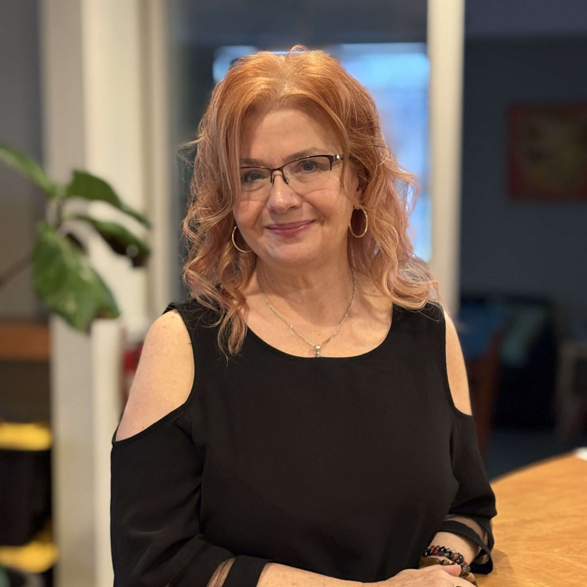 A woman with wavy red hair and glasses, wearing a black cold-shoulder top and hoop earrings, stands indoors smiling at the camera. A large green plant and blurred background furniture are visible.