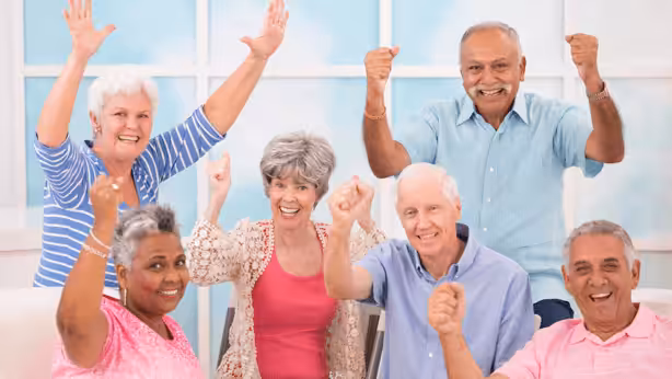 A group of six smiling older adults sit together indoors, raising their fists and arms in celebration or excitement, with a bright, cheerful background behind them.