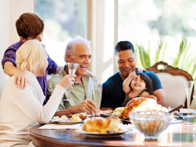 A family of five, including adults and children, sit around a dining table, smiling and enjoying a meal together. A roasted turkey and other dishes are on the table, creating a warm, festive atmosphere.