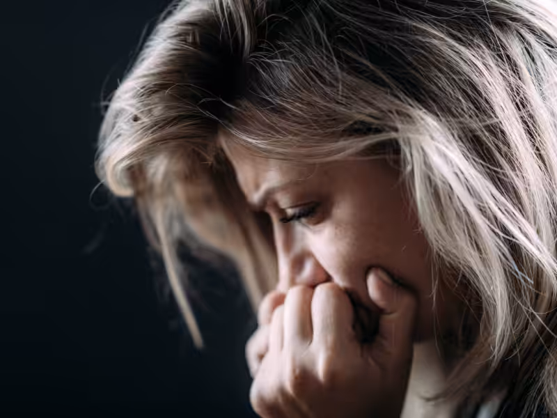 A woman with blonde hair appears upset or distressed, holding her hand to her mouth and looking down with a pained expression against a dark background.