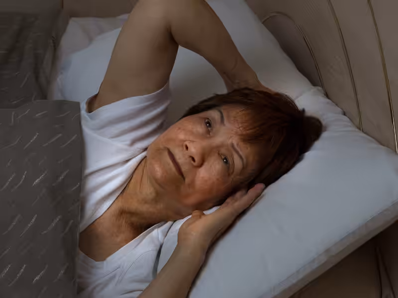 An older woman lies in bed on her back, looking concerned or unable to sleep, with one arm resting beneath her head and the other on top of her head. She is covered with a grey patterned blanket.