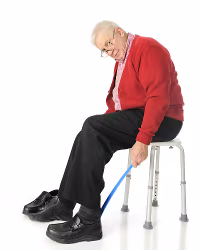 An elderly man sitting on a stool uses a long-handled shoehorn to put on his black shoes. He is wearing glasses, a red sweater, black pants, and gray socks, and appears focused on his task.