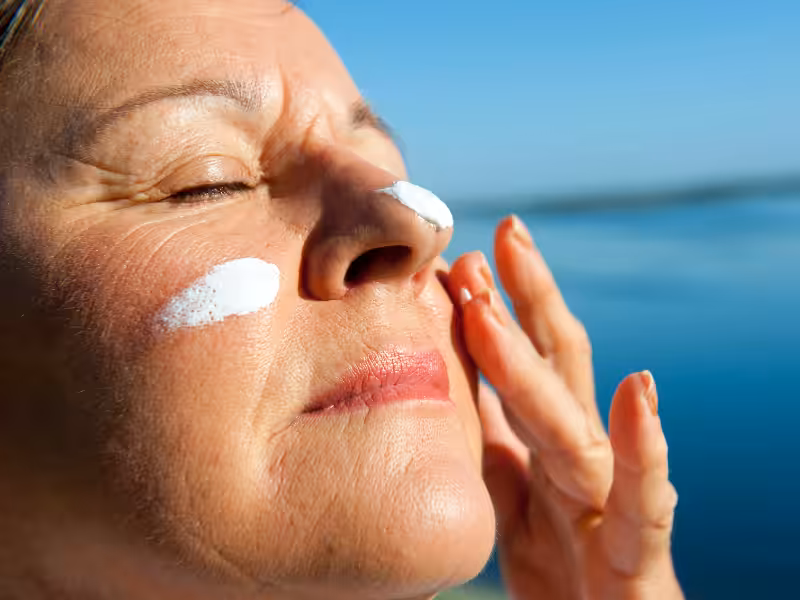 A person with closed eyes applies sunscreen to their face, with visible streaks of white lotion on their cheeks and nose, against a bright blue sky and water background.