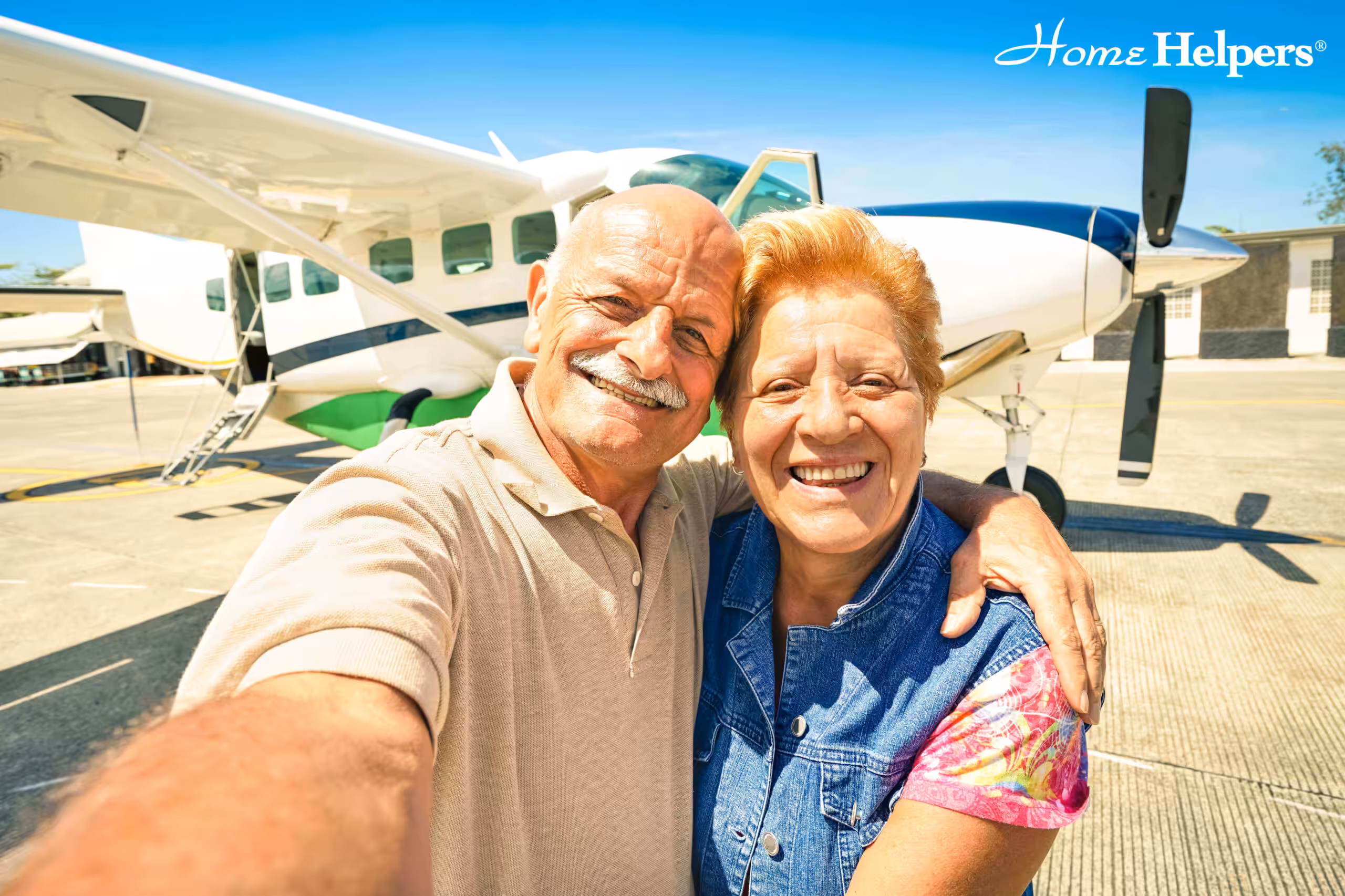 Smiling older couple taking a selfie together in front of a small airplane on a sunny day, with the Home Helpers logo in the top right corner.