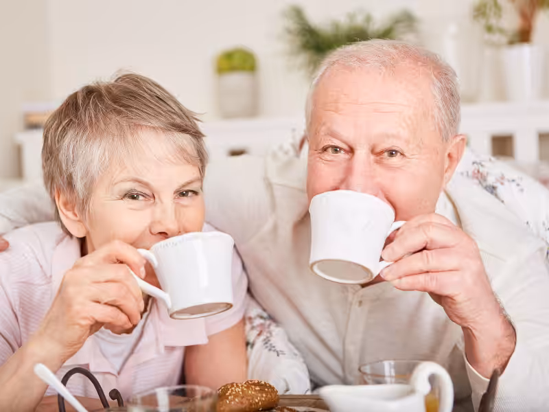 An older woman and man sit close together, smiling and drinking from white mugs at a table set with food, suggesting a warm and cheerful moment.