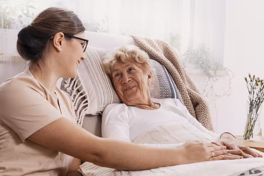 A young woman sits beside an elderly woman lying in bed, holding her hand and smiling. The elderly woman looks happy and comfortable, with a cozy blanket and pillows around her in a bright, softly lit room.