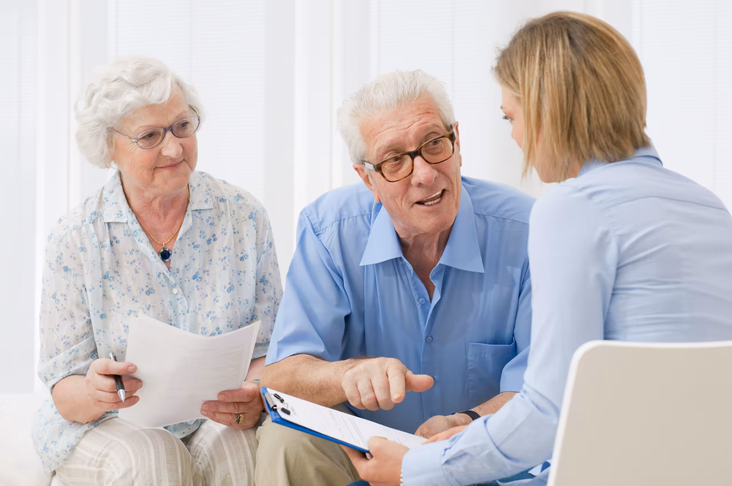 An elderly couple sits and reviews documents with a younger woman holding a clipboard, engaging in a discussion in a bright, indoor setting.