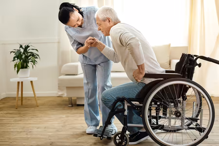 A nurse in light blue scrubs helps an elderly man in a wheelchair stand up, holding his hand in a bright, cozy living room with wooden floors and a potted plant in the background.