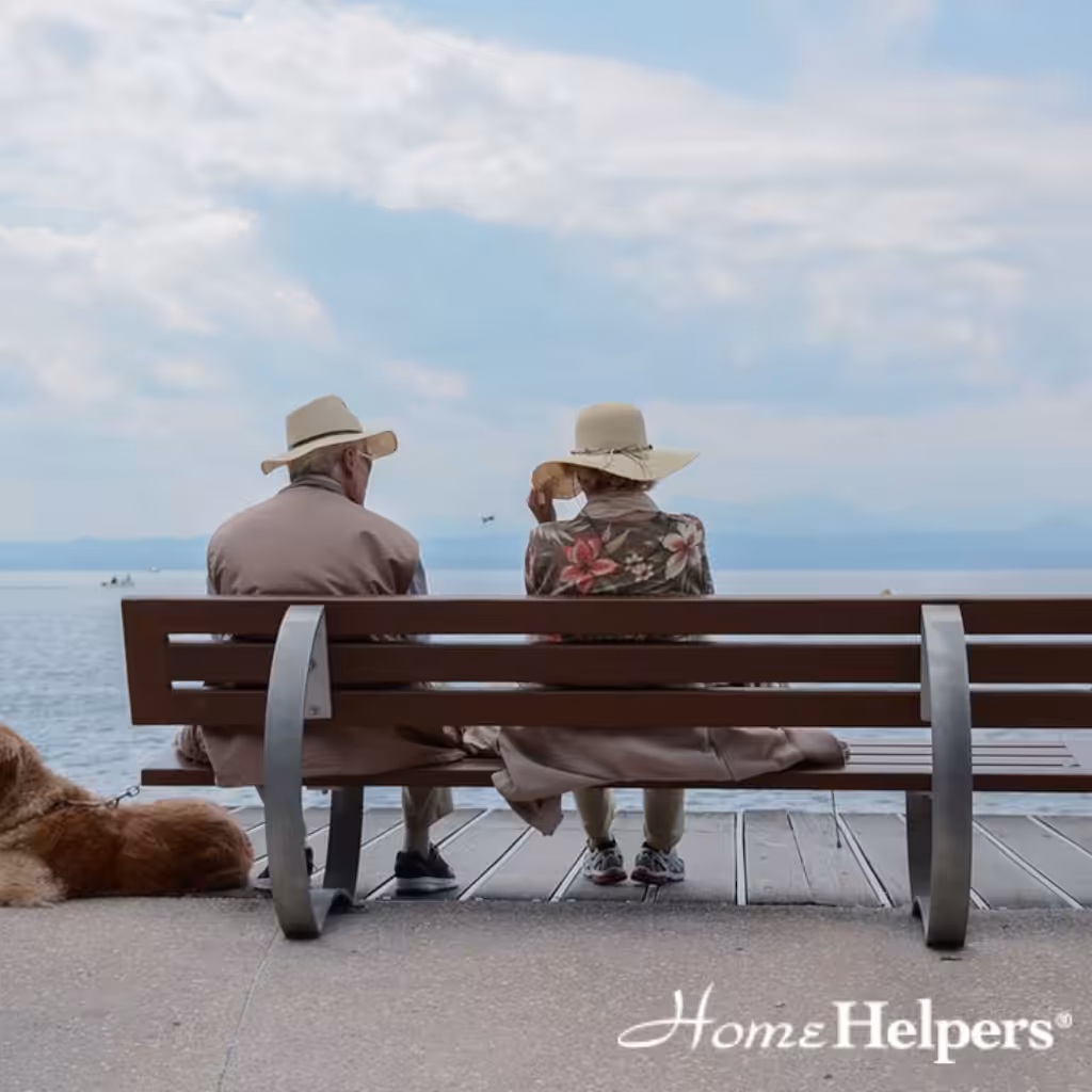 Two older adults wearing hats sit on a bench by the water, facing the sea. A dog lies beside them. The sky is partly cloudy. “Home Helpers” is written in the lower right corner.