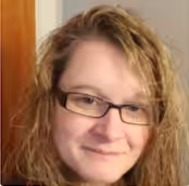 A person with wavy, light brown hair and glasses is smiling slightly, looking at the camera. They are indoors, with a wooden door and a light-colored wall in the background.