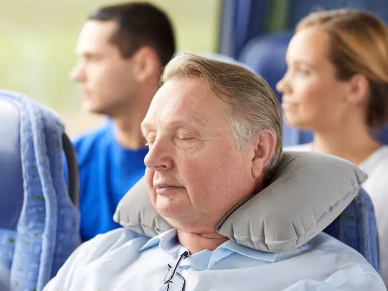 An older man with a neck pillow sleeps in a bus seat, while a younger man and woman sit behind him looking out the window. All appear relaxed and comfortable during the journey.